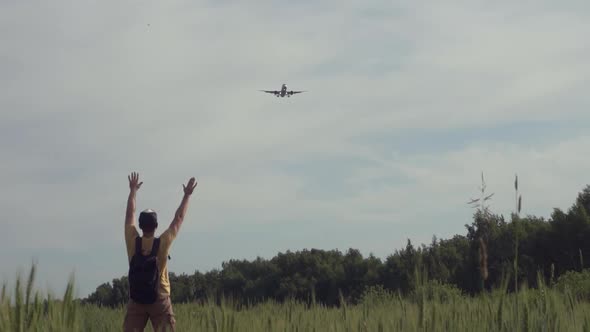 Happy Young Man Joyfully Greets an Airplane Flying in the Sky in a Wheat Field in the Summer at