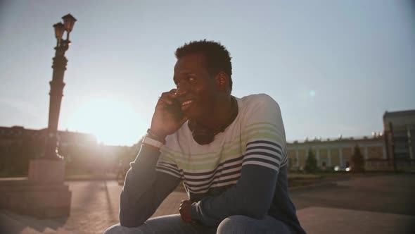 A Young Black Student in Stylish Clothes Sits and Talks on a Smartphone in the City