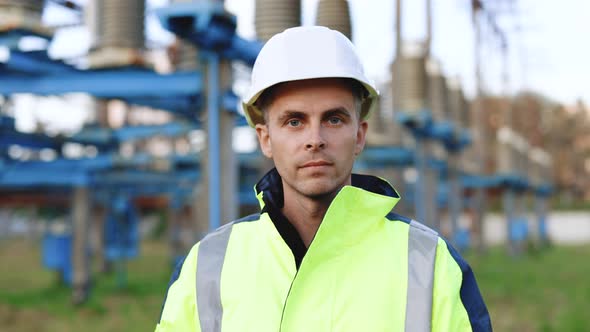 Caucasian Adult Engineer in Helmet Look at Camera at High Voltage Power Station Outside