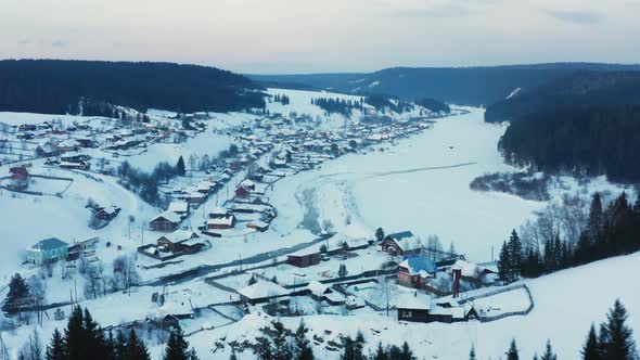 Aerial View of a Village with Wooden Houses on the Bank of a Frozen River in Winter