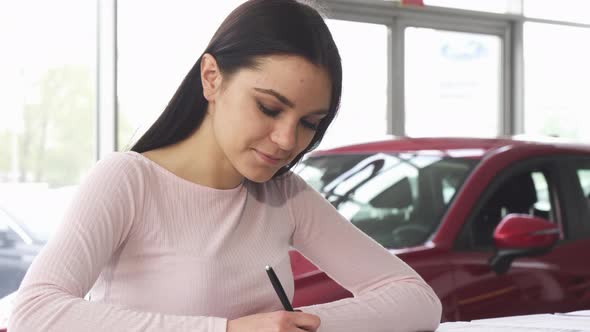 Beautiful Young Woman Signing Papers at the Dealership Showroom