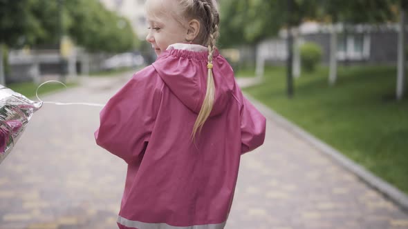 Back View of Cheerful Blond Girl with Balloon Riding Scooter. Portrait of Caucasian Kid Enjoying