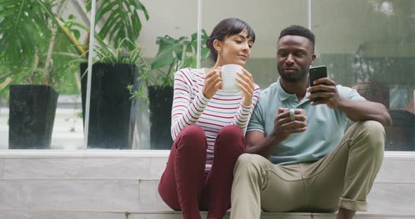 Happy diverse couple wearing blouse and shirt talking and drinking coffee in garden