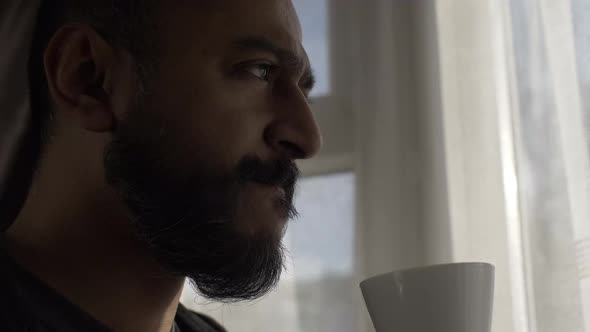 Adult Male With Beard Drinking From Cup Looking Out Through Curtains In Morning. Close Up, Locked Of