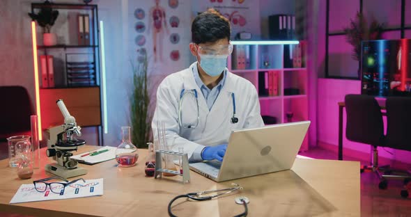 Man Scientist in Protective Mask Working on Laptop During Overtime Work in Evening Laboratory