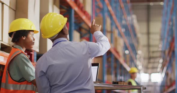 African american male and female workers wearing helmet and using laptop in warehouse