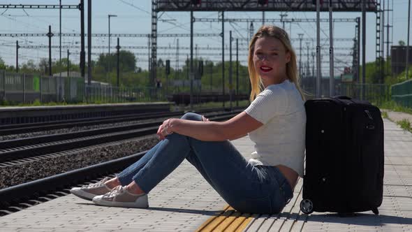 A Young Beautiful Woman Sits Next To a Suitcase on a Train Station Platform and Smiles at the Camera