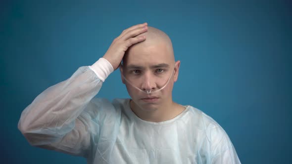 A Bald Young Man with Oncology Sadly Looks at the Camera on a Blue Background. The Patient Touches