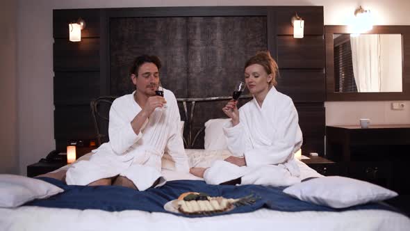 Beautiful Couple in Bathrobes Drinking Alcohol Sitting in Bed in Modern Hotel Room