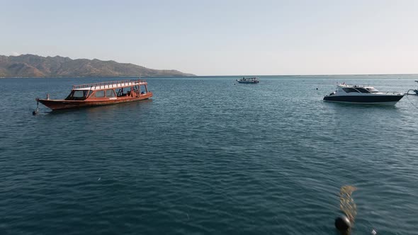 Aerial flyover boats and yachts on blue ocean water during sunny day and Lombok island in background