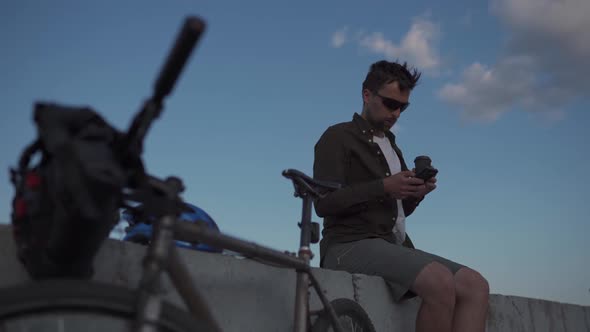Man Cyclist Resting Overlooking Sea Drinking Coffee and Using Smartphone While Sitting on Embankment