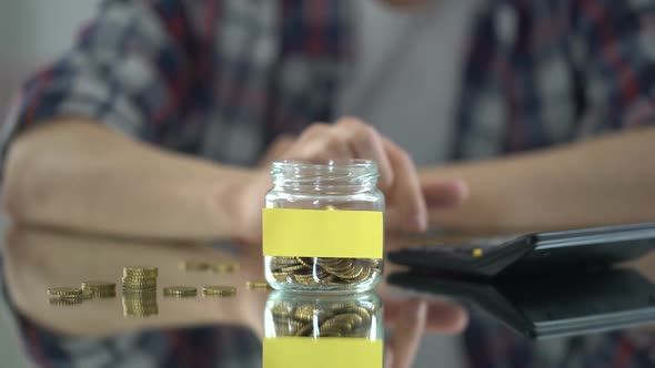 Man Counting Expenses, Putting Money Into Glass Jar With Empty Sticker for Note