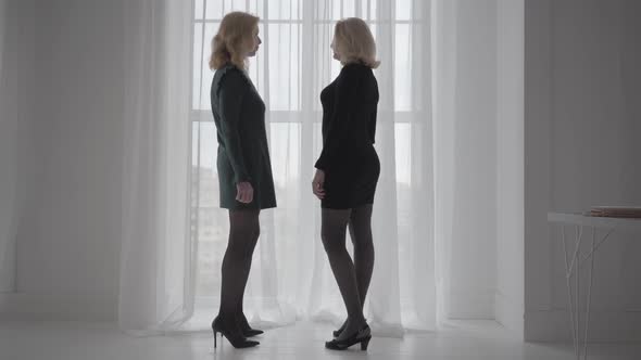 Two Mature Blonde Caucasian Women in Short Elegant Dresses Standing Next To the Window and Talking