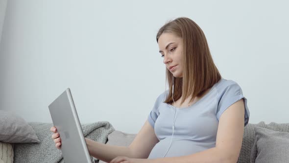 A Pregnant Woman in Home Clothes is Sitting on a Sofa and Using a Laptop