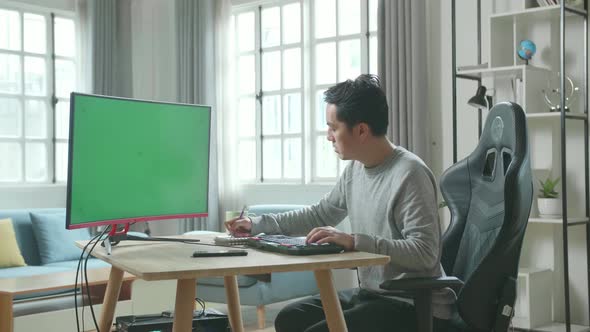 Asian Man Looking At Green Screen Desktop Computer And Writing In Notebook While Working At Home