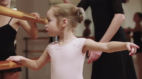 Two Ballerina Girls Training in the Studio Near the Stand with Help of Their Female Trainer