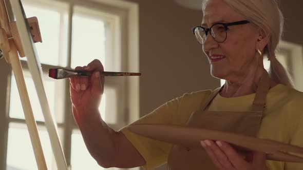 Elderly Woman Artist Paints a Picture on Canvas While Standing in a Sunny Workshop During Sunset