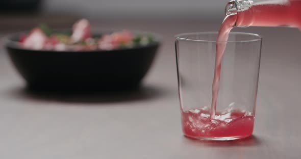 Slow Motion Pour Pink Fizzy Drink in a Tumbler Glass on Walnut Table with Salad on Background