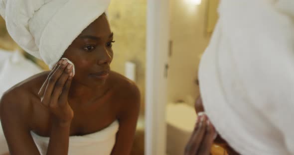 Smiling african american woman with towel watching in mirror and using pad on her face in bathroom