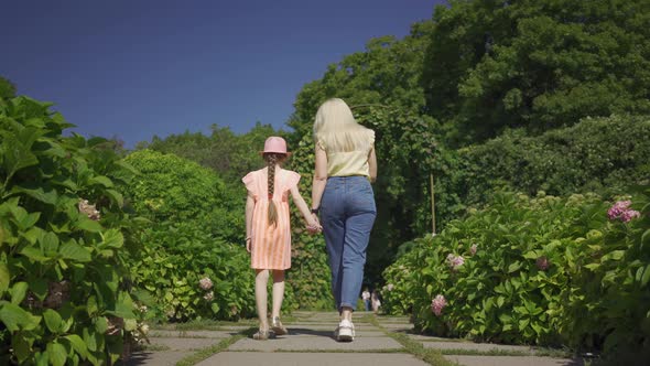 Back View of Cute Blond Mother Walking with Little Adorable Daughter in the Summer Garden Holding