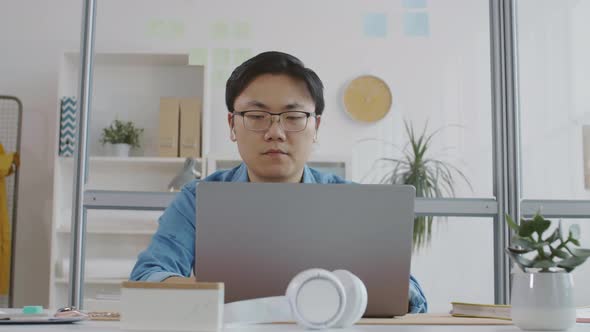 Asian Office Worker Typing on Laptop and Checking Notes in Notepad
