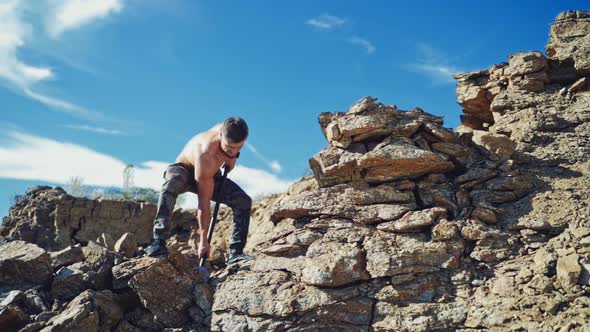 Athletic man exercises with a big hammer in canyon. Muscular man standing on rock and breaking stone