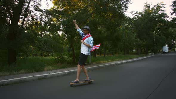 Young Man is Hanging Flag of USA on His Neck and Raising Fist Up While Skateboarding Along Deserted