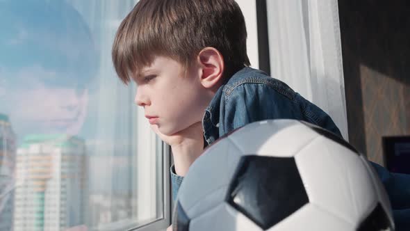 Caucasian Boy on a Home Quarantine Looking Out the Window a Bored Boy Holds a Soccer Ball