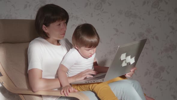 Mother and Her Toddler Boy Sit Together on Chair with Laptop. Woman Tries To Remote Work, but Kid Is