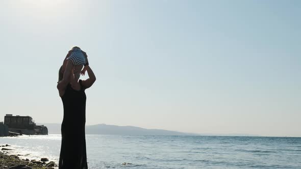 Sexy Mother in Black Long Dress on Seashore or Ocean Throws Her Daughter Up. Child in Striped Suit