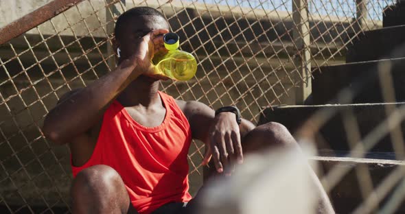 Tired african american man sitting, drinking from water bottle, taking break in exercise outdoors