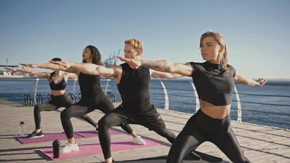 Group of Multicultural Yogi People Making Warrior Pose Outdoors Training Yoga Together on Pier in