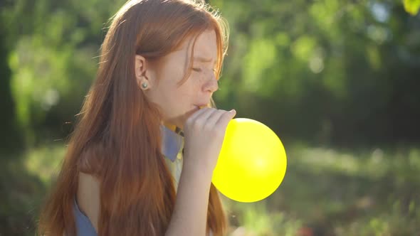 Side View Carefree Teenage Redhead Girl Blowing Yellow Balloon Sitting in Sunrays in Spring Summer