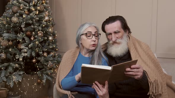Senior Couple Spending Happy Time at Home Reading a Book Together with a Decorated Christmas Tree