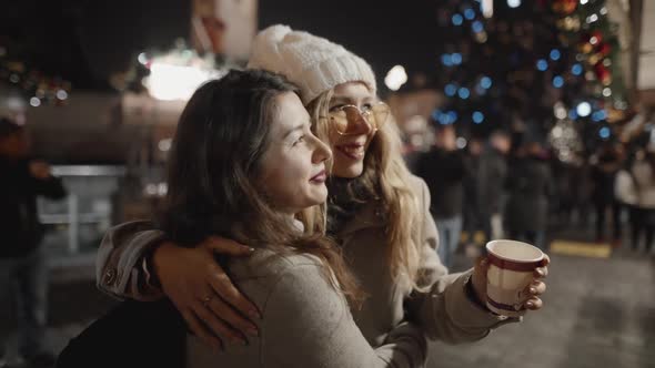 Portrait of Two Pretty Woman Hugging in a Middle of Christmas Celebration in European Town