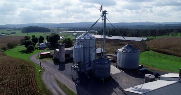 Aerial push in over cornfields to grain silos and rising up over with view of flying American flag a