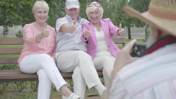 Senior Man in a Hat Makes a Photo of His Friends Sitting on a Bench in the Park