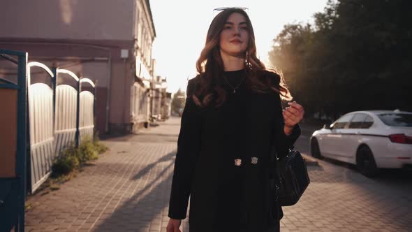 A Beautiful Young Girl in Stylish Black Clothes Goes Through the City at Sunset and Puts Sunglasses