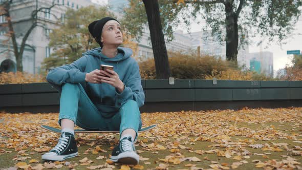 Young Caucasian Hipster Woman Using Phone in the Park Covered in Autumn Leaves While Sitting on