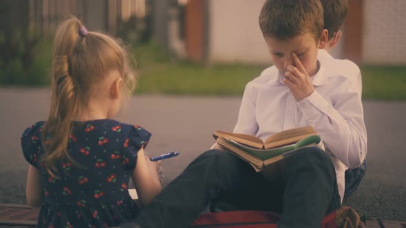 Little Girl with Pencil Talks To Boy Reading Book on Street