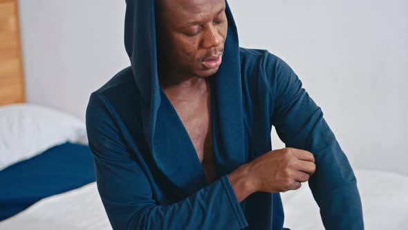 Happy African Guy Sitting on the Bed Straightening His Hoodie Smiling at the Camera