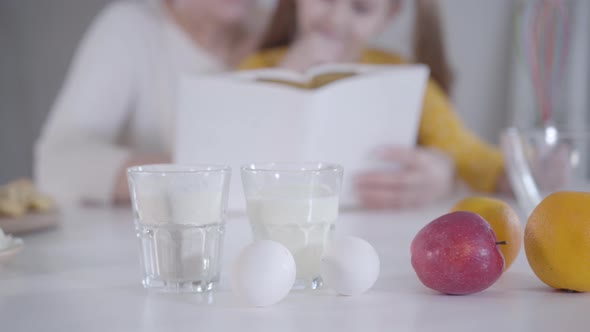 Close-up of Milk, Eggs, Flour and Fruits on the Table with Blurred People Reading Recipe at the