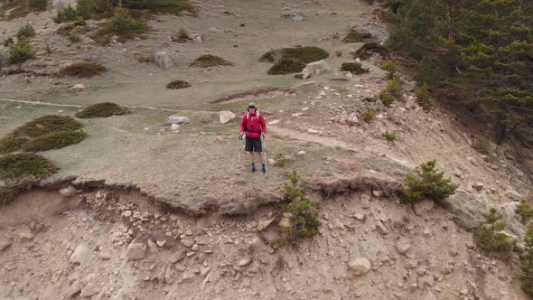 Aerial View of a Man in a Cap and Shorts and with a Backpack Walks with Trekking Poles Along a