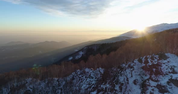 Backward Aerial Top View Over Winter Snowy Mountain and Woods Forest at Sunset or Sunrise