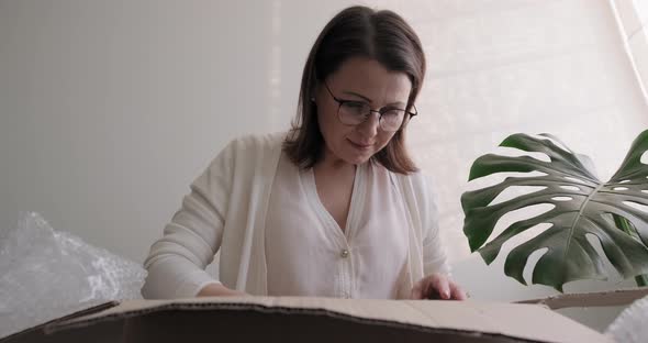 Mature Woman Unpacks Cardboard Box on Table at Home, in Office