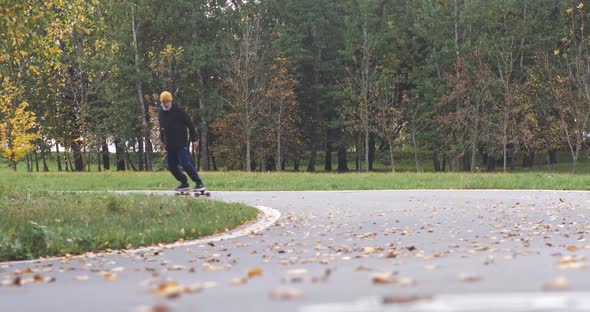 Wellness, Adult Gray-haired Man Rides a Skateboard on a Path in a Forest Park, Autumn Time of the