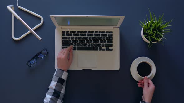 Creative Girl in a Gray Shirt Is Typing on a Laptop While Sitting at the Table and Drinks Coffee