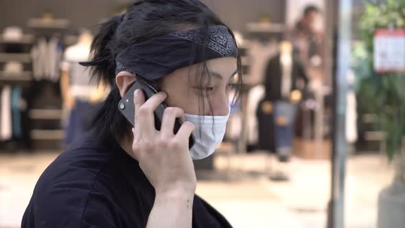 Young Asian Man in a Medical Mask Talking on a Smartphone in a Shopping Mall