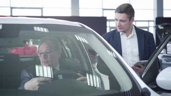 Confident Young Caucasian Car Dealer Consulting Elderly Customer Sitting Inside the Vehicle