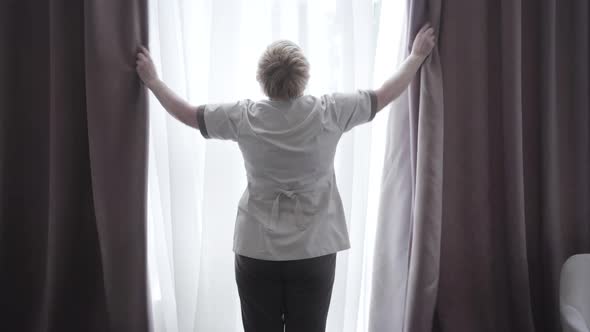 Back View of Professional Hotel Maid Opening Curtains in Hotel Room. Diligent Caucasian Adult Woman